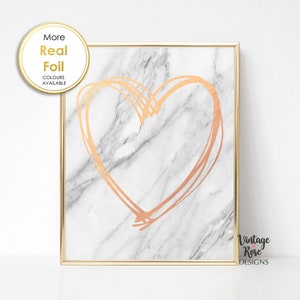 May include: A gold foil heart design on a white marble background. The print is framed in a gold frame. The text "More Real Foil" and "Colours Available" is in a circle at the top left of the image. The text "Vintage Rose Designs" is at the bottom right of the image.