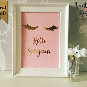May include: A white frame with a pink background and gold foil lettering that says "Hello Gorgeous". The frame has two gold foil eyelashes above the text. The frame is sitting on a table with a small vase of white flowers.