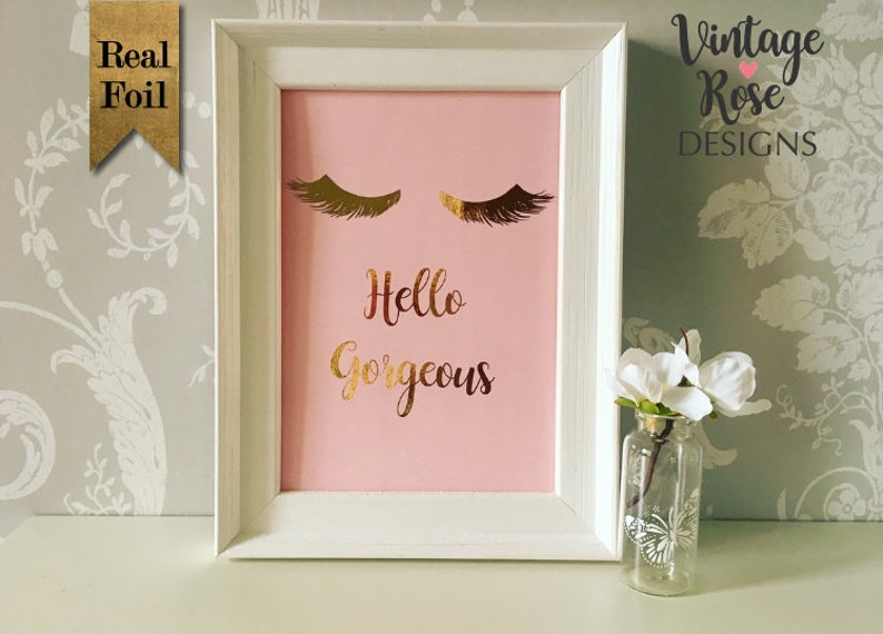 May include: A white frame with a pink background and gold foil lettering that says "Hello Gorgeous". The frame has two gold foil eyelashes above the text. The frame is sitting on a table with a small vase of white flowers.