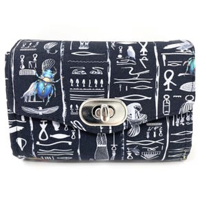 May include: A black clutch with a silver clasp, featuring a repeating pattern of Egyptian hieroglyphs and scarab beetles in white and blue. The rectangular bag has a flap closure.