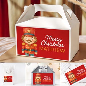 Puede incluir: Caja de regalo blanca con un panel rojo que presenta una ilustración de acuarela de un soldado cascanueces. El texto "Merry Christmas MATTHEW" se muestra en blanco y naranja. La caja tiene un asa.