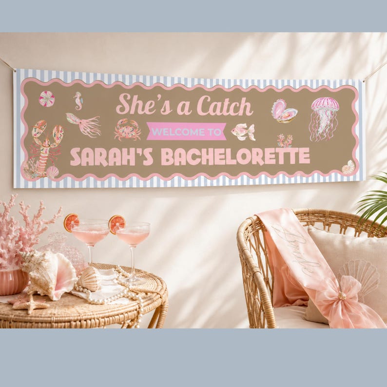 K&ouml;nnte beinhalten: Ein Banner mit dem Text "She's a Catch" und "Sarah's Bachelorette" in Rosa und Wei&szlig;. Das Banner hat einen braunen Hintergrund mit Meeresmotiven und einen gewellten Rand. Zwei Cocktails und eine Muschel stehen auf einem Tisch.