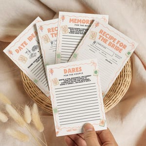 May include: A collection of light pink and white cards with various text prompts for a wedding. The cards include prompts for memories, recipes, dares, and date ideas. The cards are decorated with small illustrations and Chinese-style lanterns.
