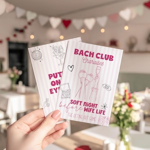 May include: Two pink and white striped cards with the text "BACH CLUB Charades" and "SOFT NIGHT before WIFE LIFE". The cards have illustrations of a disco ball, cocktails, and a heart. The cards are held in a hand.