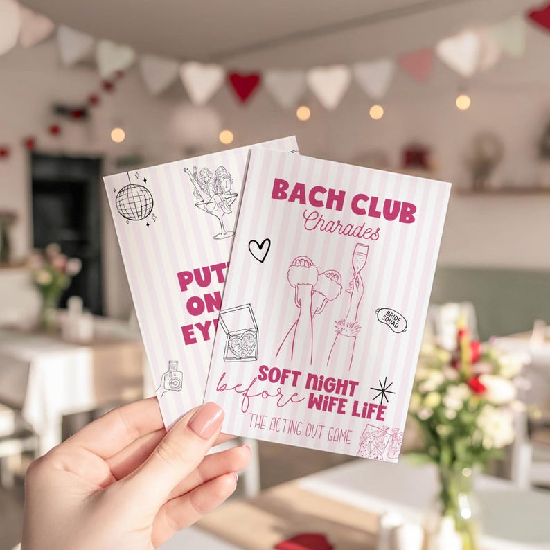 May include: Two pink and white striped cards with the text "BACH CLUB Charades" and "SOFT NIGHT before WIFE LIFE". The cards have illustrations of a disco ball, cocktails, and a heart. The cards are held in a hand.