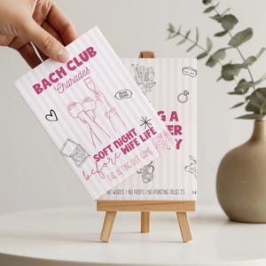 May include: A pink and white striped game card titled "BACH CLUB Charades" with illustrations and text. The card is held in front of a small wooden easel displaying a similar card. The text includes "SOFT NIGHT before WIFE LIFE" and "THE ACTING OUT GAME".