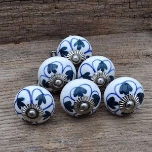 May include: Six white ceramic knobs with blue and dark green floral designs. Each knob has a silver metal centre.
