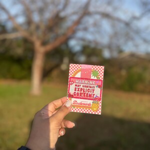 May include: A pink and white bookmark with a checkered pattern and the text "WARNING MAY CONTAIN EXPLICIT CONTENT" in black. The bookmark also features a rainbow, a heart, and a pumpkin.