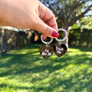 Könnte beinhalten: Zwei herzförmige Schlüsselanhänger aus schwarzem Acryl, befestigt an silbernen Metallringen. Ein Herz trägt den weißen Text "I'm Not OK" und das andere Herz den weißen Text "Trust Me" in einer stilisierten Schriftart. Die Anhänger haben eine glänzende Oberfläche.