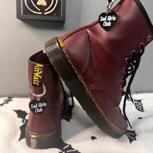 Könnte beinhalten: Ein Paar weinrote Lederstiefel mit schwarzen Schnürsenkeln und gelben Nähten. Jeder Stiefel hat einen schwarzen herzförmigen Anhänger mit der Aufschrift "Sad Girls Club". Die Stiefel haben das "AirWair"-Logo.