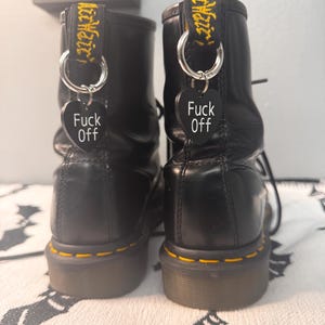 Könnte beinhalten: Paar schwarze Leder-Ankle Boots mit gelben Nähten und Marken-Zuglaschen. Jeder Stiefel hat einen silbernen Ring und einen schwarzen herzförmigen Anhänger mit dem weißen Schriftzug "Fuck Off". Die Stiefel stehen auf einer gemusterten Oberfläche.