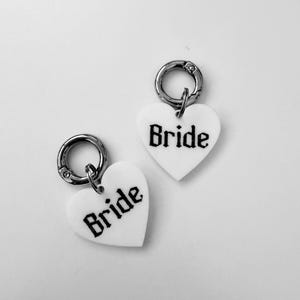 Könnte beinhalten: Zwei weiße herzförmige Anhänger mit silberfarbenen Karabinerverschlüssen aus Metall. Jeder Anhänger ist mit dem Wort Bride in einer schwarzen gotischen Schrift bedruckt. Die Anhänger sind in Design und Farbe identisch.
