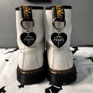 May include: White leather boots with black soles and yellow stitching. Each boot features a black heart-shaped charm with a white umbrella design, attached to a silver ring. The boots have yellow and black pull tabs.