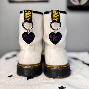 Könnte beinhalten: Zwei weiße Lederstiefel mit schwarzen Sohlen und gelben Nähten. Jeder Stiefel hat einen schwarzen herzförmigen Anhänger mit dem lila Text "It Wasn't a Phase", befestigt mit einem silbernen Ring. Die Stiefel haben schwarze und gelbe Zuglaschen.