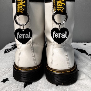 Puede incluir: Un par de botas de cuero blanco con suelas negras y costuras amarillas. Cada bota tiene un dije negro en forma de corazón que dice "feral" en letras blancas, sujeto al tirador.
