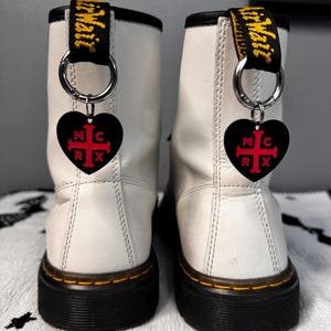 K&ouml;nnte beinhalten: Paar wei&szlig;e Lederstiefel mit schwarzen Sohlen und gelben N&auml;hten. Jeder Stiefel hat einen schwarzen herzf&ouml;rmigen Anh&auml;nger mit den Buchstaben "MCRX" und einem roten Kreuz-Design. Die Stiefel haben schwarze und gelbe Zuglaschen.