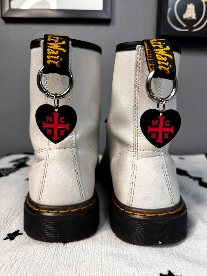 K&ouml;nnte beinhalten: Paar wei&szlig;e Lederstiefel mit schwarzen Sohlen und gelben N&auml;hten. Jeder Stiefel hat einen schwarzen herzf&ouml;rmigen Anh&auml;nger mit den Buchstaben "MCRX" und einem roten Kreuz-Design. Die Stiefel haben schwarze und gelbe Zuglaschen.