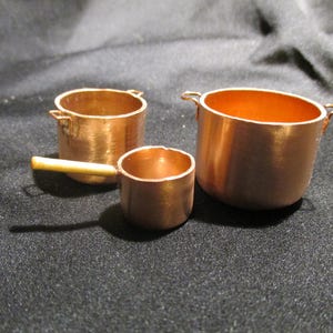 May include: Three miniature copper pots. The largest pot has two small handles. The middle pot has a handle made of wood. The smallest pot has a small handle. All three pots are made of copper and have a shiny finish.