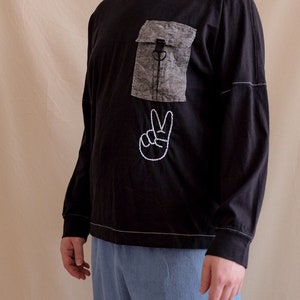 Pode incluir: Uma camisa preta de manga comprida com um bolso de remendo cinza e um sinal de paz bordado em branco.