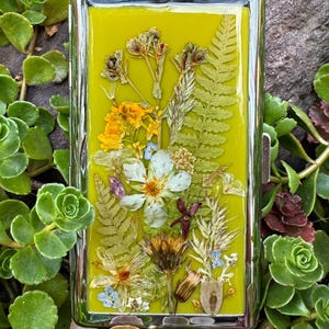 May include: A rectangular metal cigarette case with a polished silver frame and a lime green resin inlay. The surface is embedded with a variety of pressed dried flowers, including white blossoms, yellow petals, and green fern fronds.