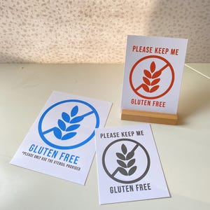 Puede incluir: Tres carteles con el texto "Gluten Free" y un símbolo de trigo tachado. Los carteles son blancos con símbolos de trigo rojo, azul y gris. El texto "Please Keep Me" está en la parte superior de cada cartel.