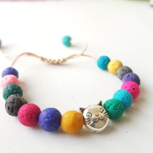 Peut inclure: Un bracelet en pierre de lave coloré avec un charme de chat argenté. Le bracelet est fait avec un cordon brun et présente des perles dans des tons de rose, violet, bleu, vert, jaune et noir.