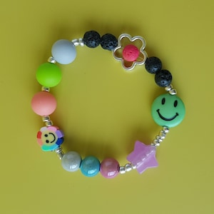 Könnte beinhalten: Ein farbenfrohes Perlenarmband mit einem Smiley-Anhänger, einem Blumenanhänger und einem Sternanhänger. Das Armband besteht aus Lava-Perlen, grauen, grünen, rosa und blauen Perlen.