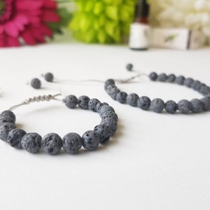 May include: Two black lava stone beaded bracelets with adjustable string closures. The bracelets are on a white surface.