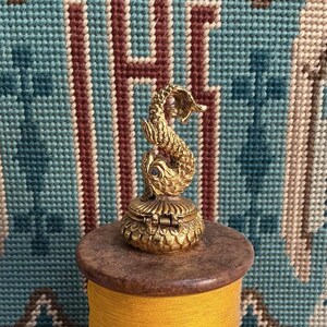 May include: A small, gold-toned metal trinket box featuring a sculpted fish figure on the lid. The fish has textured scales and a single dark blue stone set as an eye. The base of the box is circular with a hinged lid and a detailed scale pattern.