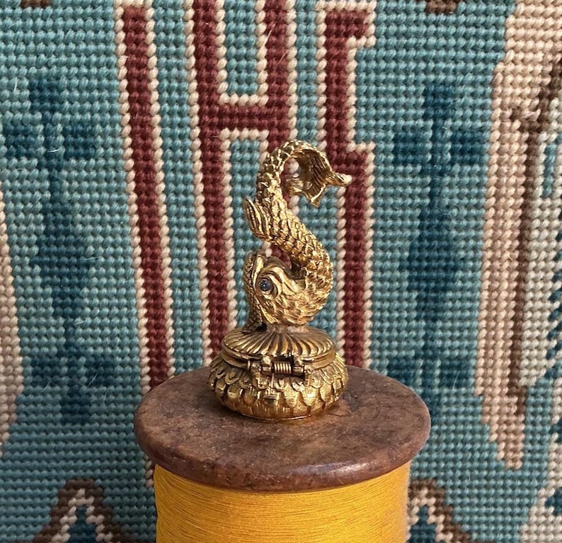 May include: A small, gold-toned metal trinket box featuring a sculpted fish figure on the lid. The fish has textured scales and a single dark blue stone set as an eye. The base of the box is circular with a hinged lid and a detailed scale pattern.