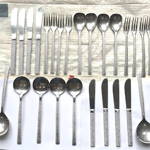 May include: A set of vintage cutlery, including knives, forks, and spoons. The stainless steel utensils have textured handles and are arranged on a white surface. The spoons have a rounded bowl shape.