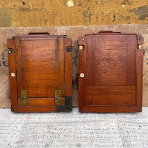 Puede incluir: Dos respaldos de cámara de madera antiguos con herrajes de latón. Los respaldos rectangulares son de un color marrón rojizo intenso, con la veta de la madera visible. Cada uno tiene un pequeño dial blanco y bisagras de latón. Probablemente son para una cámara de gran formato.