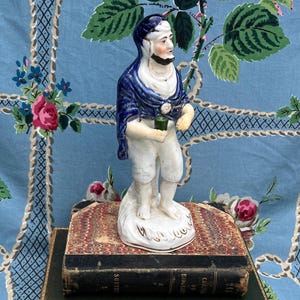 Puede incluir: Figura de porcelana vintage de un hombre con chal azul y pantalones blancos, sosteniendo un pequeño objeto verde. La figura está sobre una base blanca con detalles dorados, colocada sobre libros antiguos. Fondo de tela azul con estampado floral.