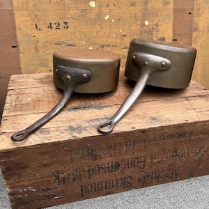 May include: Two antique copper saucepans with long, riveted handles. The pans have a rounded shape and a dark, aged patina. They rest on a weathered wooden crate, suggesting a vintage or rustic aesthetic. The handles are made of a lighter metal.