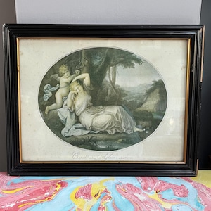 Könnte beinhalten: Gerahmtes Kunstwerk mit einer klassischen Szene mit einer Frau und einem Cherub. Das Kunstwerk befindet sich in einem ovalen Rahmen, der sich in einem schwarzen rechteckigen Rahmen mit goldenem Rand befindet. Das Kunstwerk ist signiert "Cupid and Psyche" von W. L. Laurence.