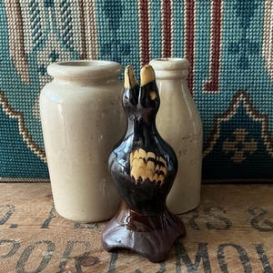 May include: A dark brown and black ceramic duck figurine with yellow detailing, positioned between two beige ceramic jars. The duck features a detailed feather design. The background includes a patterned textile and a wooden surface.