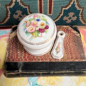May include: A white ceramic knob and matching pull with floral designs in pink, yellow, and blue. The knob and pull have gold trim and sit on a vintage book with marbled paper.