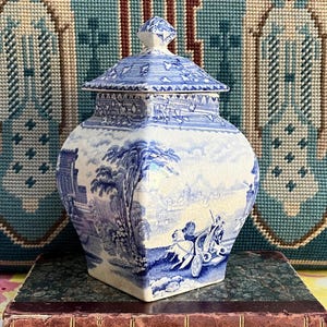 May include: A square ceramic jar with a lid, featuring a blue and white transferware pattern depicting a horse-drawn carriage, trees, and architectural structures. The surface has a visible crackle glaze finish and a pointed finial on the lid.