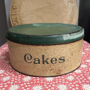 May include: A round, vintage-style metal storage tin with a cream-colored base featuring a speckled, distressed finish and the word "Cakes" printed in dark lettering. The tin has a solid, dark green lid with visible signs of wear.