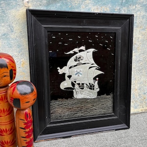 Puede incluir: Un espejo con marco negro con una silueta de barco plateada sobre un fondo negro. El barco tiene una vela blanca y está rodeado de pequeños pájaros blancos.