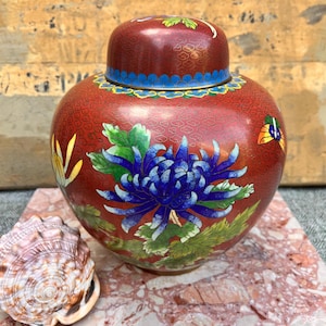 May include: A round red cloisonné jar with a matching lid, featuring a prominent blue chrysanthemum flower, green leaves, and a small butterfly. The surface has a fine gold wire pattern throughout, with blue and yellow decorative border accents.