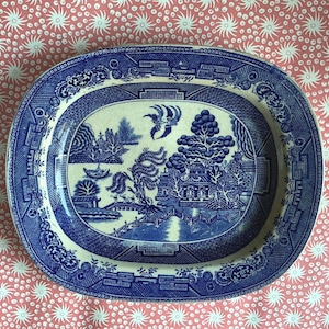 May include: An oval ceramic platter featuring a blue and white willow pattern. The central design depicts a landscape with a bridge, pagoda, trees, and birds. The wide rim is decorated with a geometric border pattern in matching blue and white.