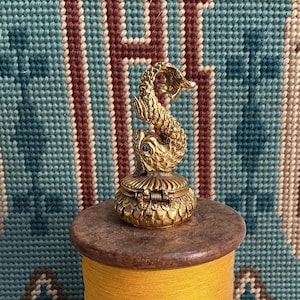 May include: A small, gold-toned metal trinket box featuring a sculpted dolphin figure on the lid. The box has a textured, scale-like pattern on its base and a hinged closure. The dolphin is depicted in a curved, upright position with fine detailing.