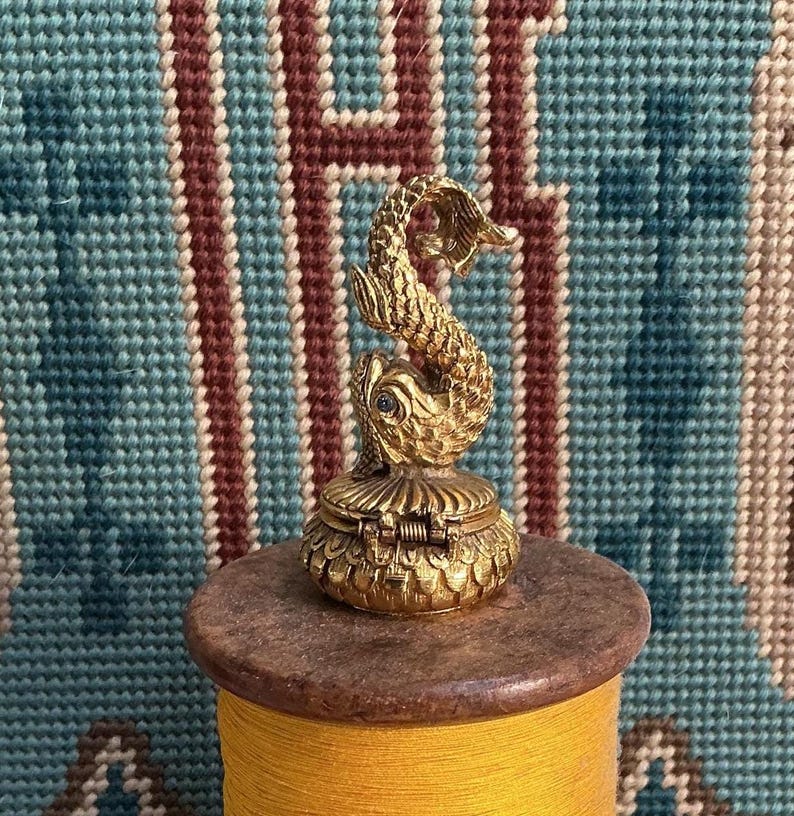 May include: A small, gold-toned metal trinket box featuring a sculpted dolphin figure on the lid. The box has a textured, scale-like pattern on its base and a hinged closure. The dolphin is depicted in a curved, upright position with fine detailing.