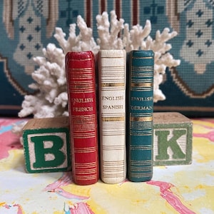 May include: Three miniature books with gold lettering and decorative spines. The books are red, white, and teal. The spines read "English French", "English Spanish", and "English German". Wooden blocks with the letters "B" and "K" are in the foreground.
