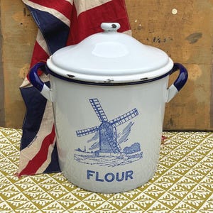 Könnte beinhalten: Ein weißer Mehlbehälter aus Emaille mit Deckel, zwei dunkelblauen Seitengriffen und einer blauen Windmühlengrafik auf der Vorderseite über dem Wort FLOUR. Der Behälter hat einen dunkelblauen Rand und weist Gebrauchsspuren an den Kanten auf.