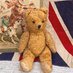 Könnte beinhalten: Ein alter Teddybär aus goldbraunem Mohair mit einer gestickten schwarzen Nase und dunklen Augen. Der Bär hat abgenutztes, strukturiertes Fell, abgerundete Ohren und helle Stoffpfoten. Er sitzt in aufrechter Position mit beweglichen Gliedmaßen.