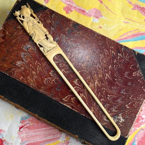 Pu&ograve; includere: Forcina per capelli color avorio con una figura scolpita che tiene un fiore di loto. L'accessorio per capelli &egrave; appoggiato su un libro d'epoca con un motivo marmorizzato e un dorso in pelle nera.