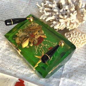 May include: A rectangular green resin paperweight containing embedded seashells, a small crab, and a seahorse. Two black and gold metal handles are attached to the top surface. The object is positioned near a white coral specimen.