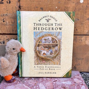 Puede incluir: Un libro pop-up tridimensional titulado Brambly Hedge Through The Hedgerow de Jill Barklem con una cubierta de color crema que presenta una ilustración circular de una casa de ratones dentro de un seto, junto a un pequeño pato de peluche con pico naranja.
